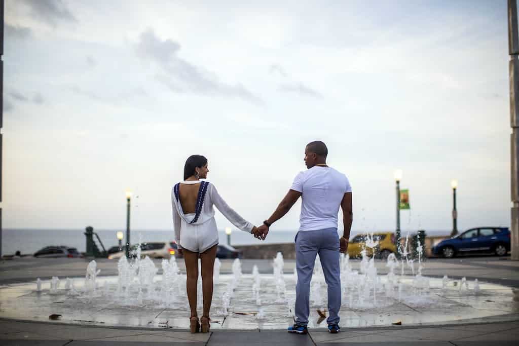 Back view full body positive African American couple in stylish summer wear holding hands and looking at each other while standing near fountain on seafront