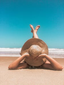 A woman in a straw hat relaxes on a sunny beach, enjoying the clear blue sky and ocean waves.