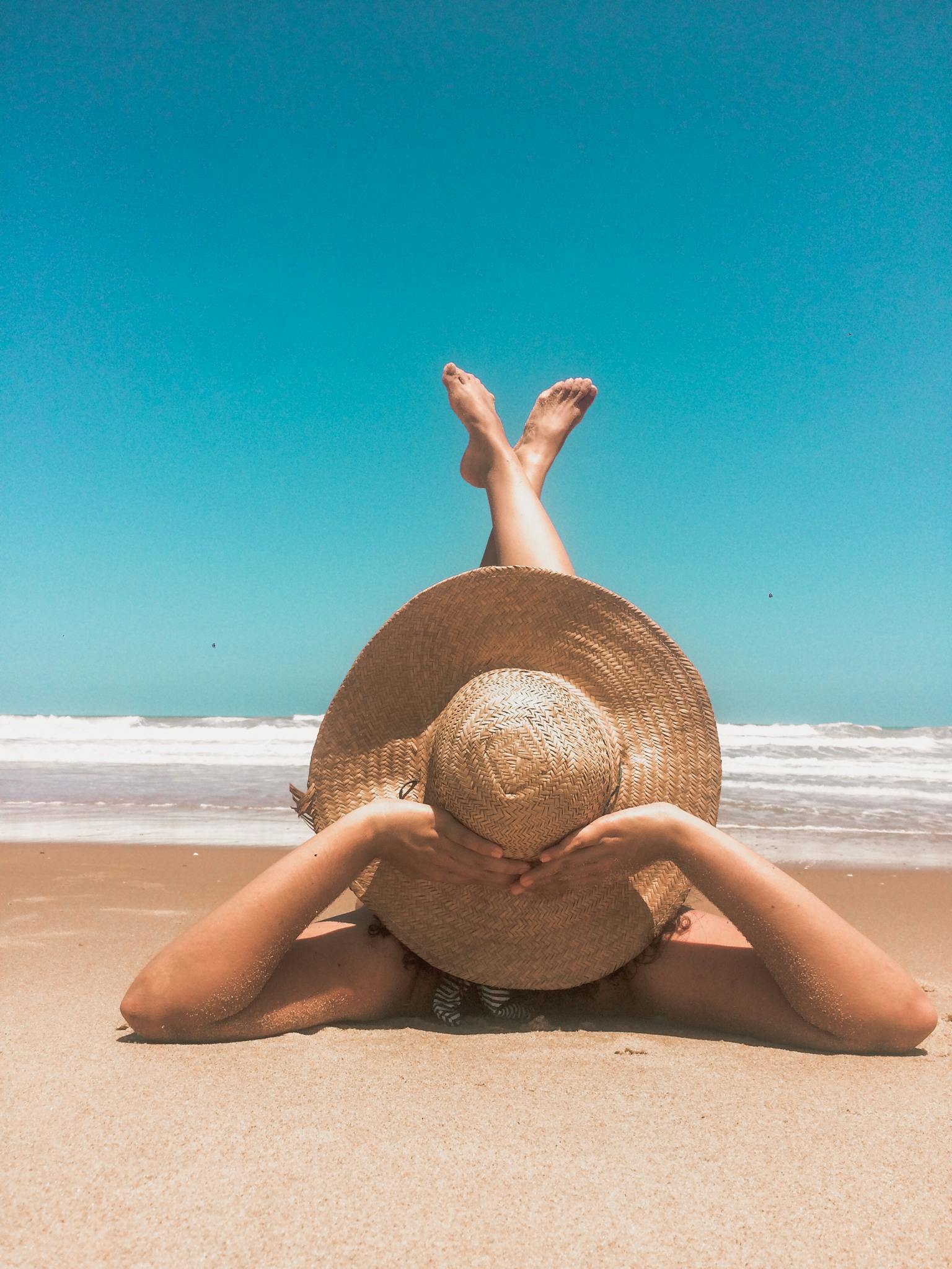 A woman in a straw hat relaxes on a sunny beach, enjoying the clear blue sky and ocean waves.