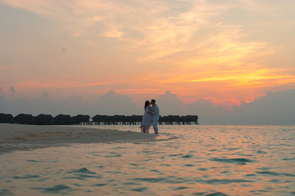 A romantic couple embraces on a tropical beach during a stunning sunset, with overwater bungalows in the background.