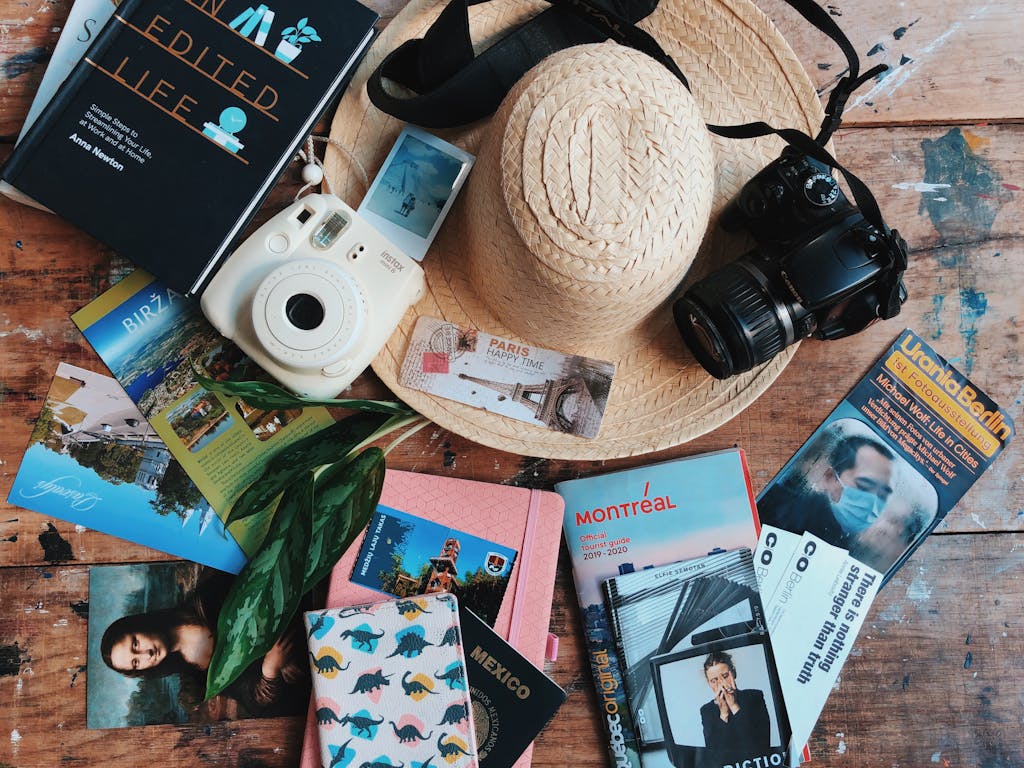 Flat lay of travel accessories including a straw hat, cameras, and guidebooks on a wooden table.